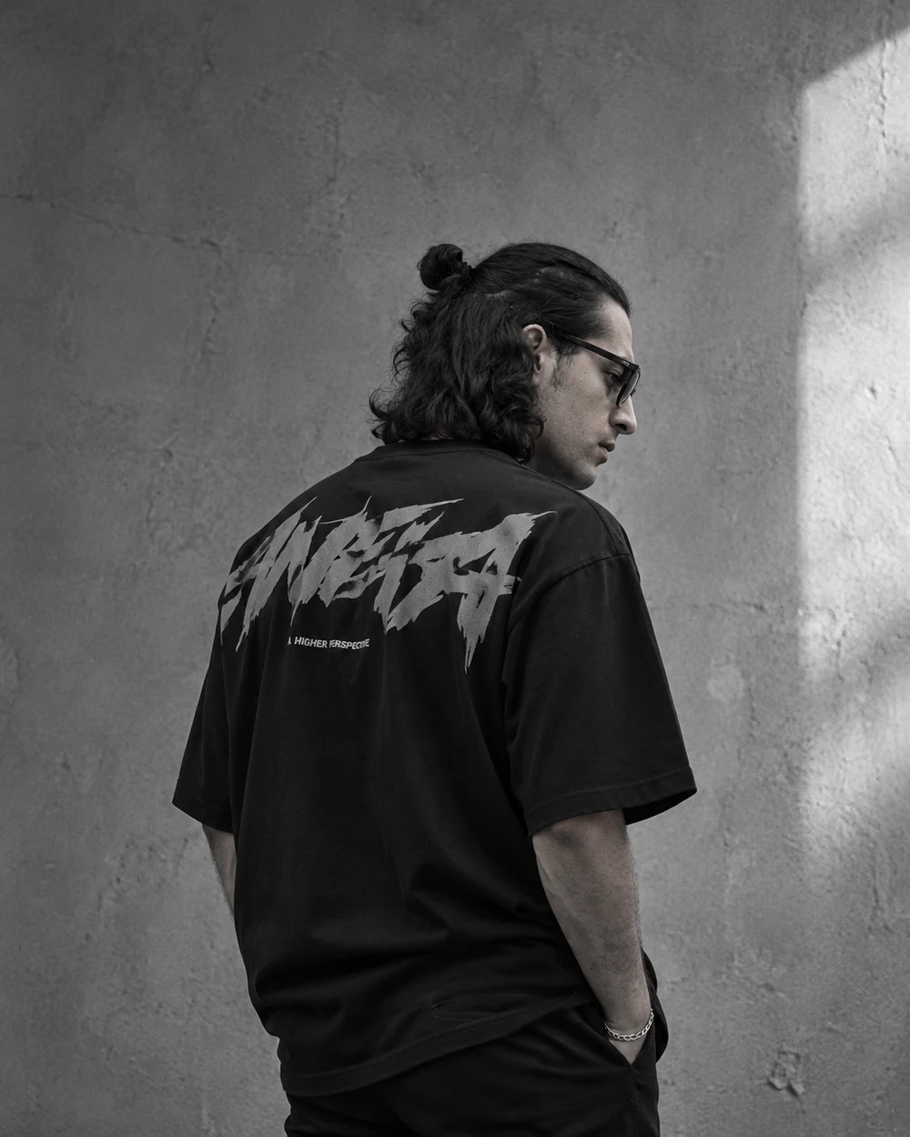 Monochrome portrait of Amir Khaldari from behind in a black t-shirt, looking down, against a concrete wall